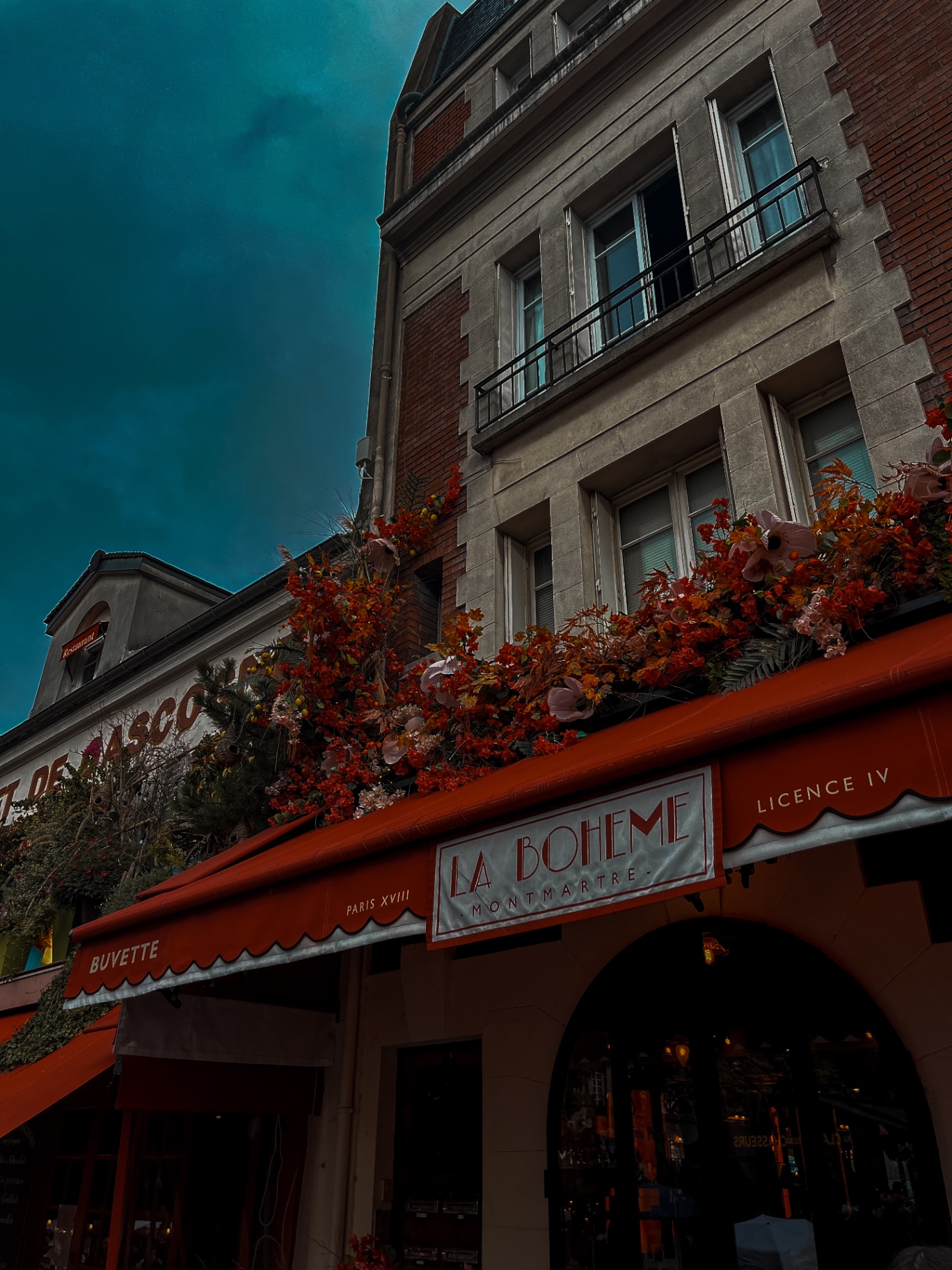 Toldo vermelho do bistrô La Bohème em Montmartre, Paris, com flores de outono e céu turquesa carregado